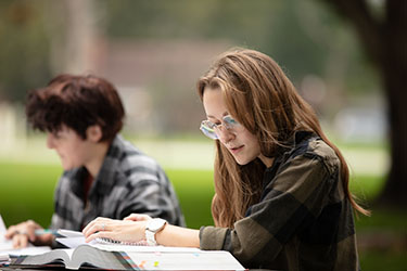 Two students sit at a picnic table outside and read books that are open on the table in front of them.